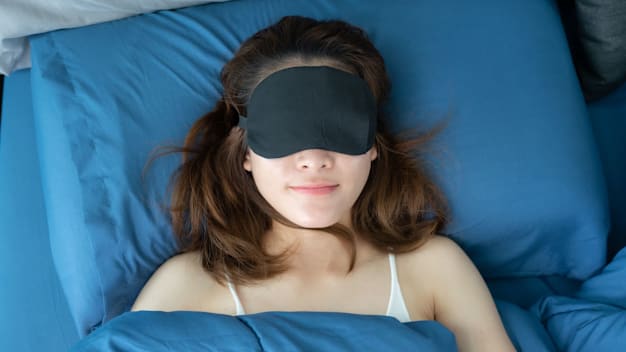 person sleeping on back while wearing sleep mask, blue pillow cover and sheets on bed